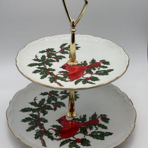 May include: Two-tiered serving tray with a gold handle and accents. Each white plate features a red cardinal bird and green holly design with gold trim. The serving tray is perfect for displaying desserts or appetizers.