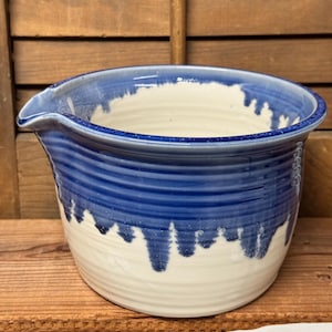 May include: A ceramic mixing bowl with a pouring spout, featuring a cream-colored base and a deep blue glazed upper section that drips down the sides. The exterior surface has horizontal ridges throughout, and the interior is smooth and cream-colored.