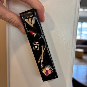 May include: Black rectangular magnet with gold embellishments. Features a gold letter "V", a red lipstick, a perfume bottle, a small square with a gem, two gold paint brushes, and a makeup palette with colorful eyeshadows.