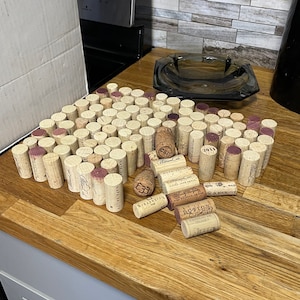 Può includere: Una grande collezione di tappi di vino usati, molti con etichette, disposti in forma rettangolare su una superficie di legno. Alcuni tappi sono sparsi intorno al gruppo principale.