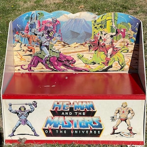 Op de afbeelding: Vintage speelgoedkist met He-Man and the Masters of the Universe-personages. De kist heeft een rode binnenkant en een witte buitenkant met kleurrijke illustraties van de personages. De voorkant van de kist toont het logo van de show en karakterillustraties.