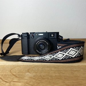 Op de afbeelding: Zwarte digitale camera met een patroon camerariem. De riem heeft een herhalend geometrisch ontwerp in wit, bruin en zwart. De camera ligt op een houten oppervlak.