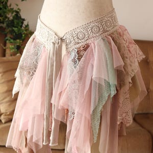 Könnte beinhalten: Ein mehrlagiger Rock im Bohème-Stil. Der Rock hat mehrere Schichten aus rosa, beige und mintgrünem Stoff mit Spitzenbesatz. Der Bund ist cremefarben mit einem dekorativen Spitzendesign.