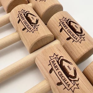May include: A collection of light-colored wooden mallets, each featuring a laser-engraved design of a crab inside a banner with the text BABY CECILIA and the year 2025. The mallets have cylindrical heads and long, smooth, rounded wooden handles.