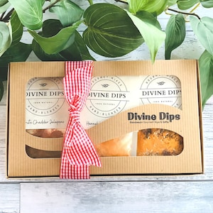 May include: A gift box with three jars of gourmet dip mixes. The jars are labeled "Divine Dips" and feature a red and white gingham ribbon. The box is made of brown cardboard and has a window to show the contents.
