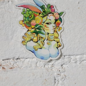 May include: A colorful sticker featuring a woman with a crown of fruit and vegetables. The woman has blue eyes and a light blue and white background.