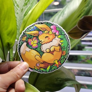 May include: A round sticker featuring a cartoon Eevee character, colored in shades of orange and cream, nestled among green leaves and colorful flowers. The character is smiling and wearing a flower crown. The sticker is held in front of a leafy background.