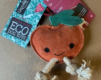 May include: An orange apple-shaped dog toy with a smiling face and rope legs. The toy has a green leaf and is attached to tags that read "GREEN&WILD'S ECO DOG TOYS" and "To: From:" on a brown surface.