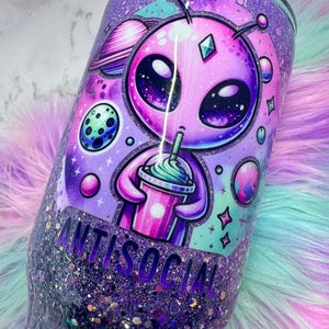 Pu&ograve; includere: Un bicchiere viola glitterato con un design alieno ed elementi spaziali. L'alieno tiene in mano una bevanda, con la scritta "ANTISOCIAL". Il bicchiere ha una base glitterata e un design vivace a tema spaziale.