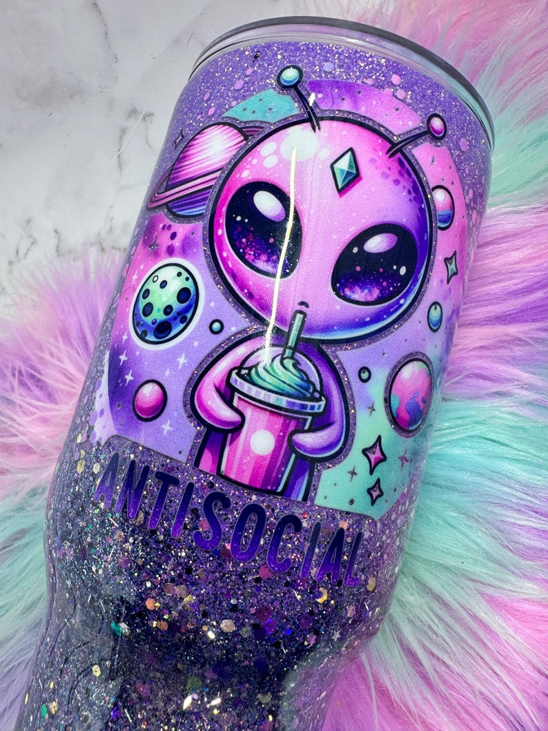 Pu&ograve; includere: Un bicchiere viola glitterato con un design alieno ed elementi spaziali. L'alieno tiene in mano una bevanda, con la scritta "ANTISOCIAL". Il bicchiere ha una base glitterata e un design vivace a tema spaziale.