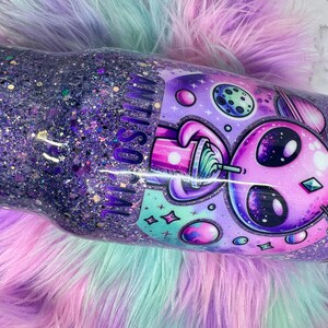 Pu&ograve; includere: Un bicchiere glitterato viola con un design alieno. Il bicchiere presenta un alieno dei cartoni animati con una bevanda, pianeti e stelle. Le parole "ANTI SOCIAL" sono visibili sul lato. Il bicchiere &egrave; su una superficie soffice e multicolore.