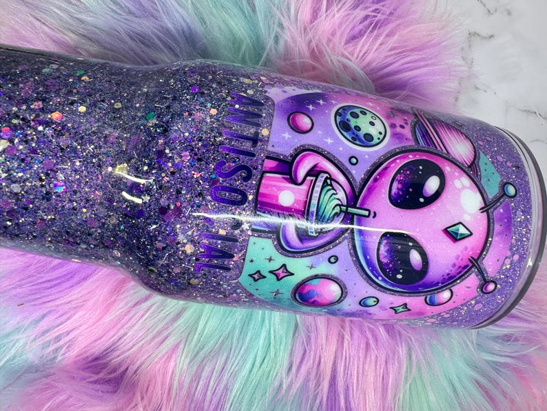 Pu&ograve; includere: Un bicchiere glitterato viola con un design alieno. Il bicchiere presenta un alieno dei cartoni animati con una bevanda, pianeti e stelle. Le parole "ANTI SOCIAL" sono visibili sul lato. Il bicchiere &egrave; su una superficie soffice e multicolore.