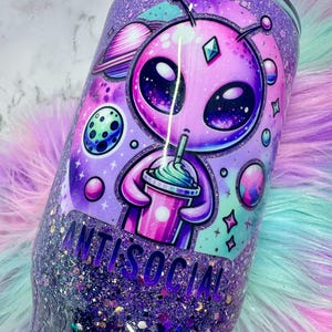 Pu&ograve; includere: Un bicchiere glitterato viola con un'illustrazione di un alieno che tiene in mano una bevanda. L'alieno &egrave; rosa con grandi occhi ed &egrave; circondato da pianeti e stelle. La parola "ANTISOCIAL" &egrave; stampata sul bicchiere.
