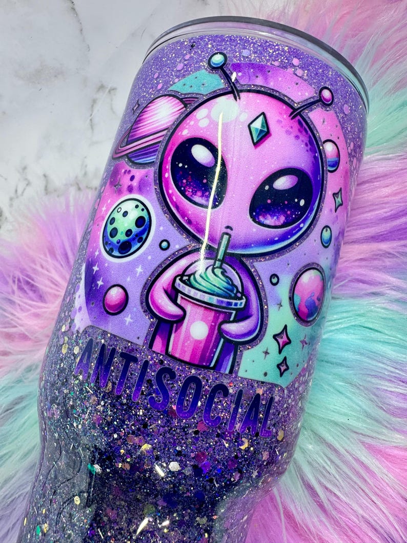 Pu&ograve; includere: Un bicchiere glitterato viola con un'illustrazione di un alieno che tiene in mano una bevanda. L'alieno &egrave; rosa con grandi occhi ed &egrave; circondato da pianeti e stelle. La parola "ANTISOCIAL" &egrave; stampata sul bicchiere.