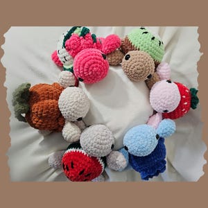 May include: A collection of handmade crochet fruit toys, including a strawberry, watermelon, pumpkin, and other colorful designs. The toys are arranged in a circular pattern on a white surface.