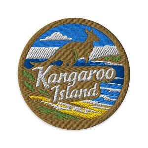 May include: A circular embroidered patch featuring a brown kangaroo silhouette against a blue sky with white clouds. The text Kangaroo Island is stitched in white across the centre, with green and yellow landscape details at the bottom.