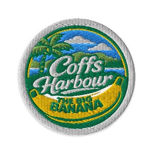 May include: A circular embroidered patch featuring a yellow banana graphic with the text Coffs Harbour and THE BIG BANANA in white and green lettering. The design includes a green palm tree, blue sky, white clouds, and green hills.