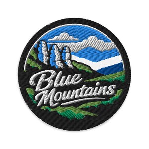 May include: A circular embroidered patch featuring a landscape of the Blue Mountains with green hills, white rock formations, and a blue sky with white clouds. The text Blue Mountains is stitched in white script across the foreground.