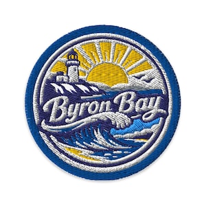 May include: Embroidered patch with a blue border featuring a lighthouse, sun, and ocean waves. The text "Byron Bay" is in white script across the centre. The design includes yellow, white, and blue colours.