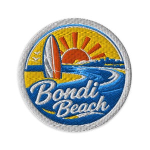May include: A circular embroidered patch featuring a yellow sky with an orange sun and a white surfboard standing on a blue beach. The text Bondi Beach is stitched in white cursive script across the lower blue section of the circular design.