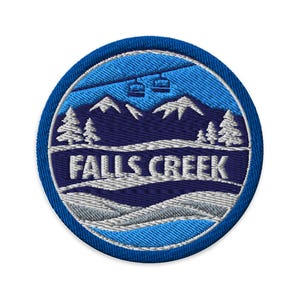 May include: A circular embroidered patch featuring a mountain landscape with snow-capped peaks, pine trees, and ski lifts against a blue sky. The text FALLS CREEK is stitched in white across the centre over a dark blue and grey snowy terrain.