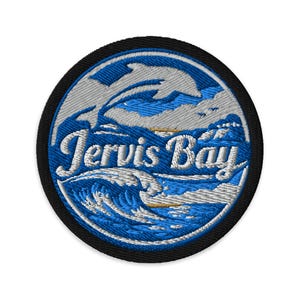 May include: Circular embroidered patch with a black border, featuring a blue and white design of waves, a dolphin, and a coastal scene. The text "Jervis Bay" is embroidered in white script.