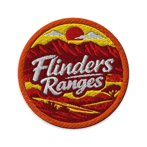 May include: A circular embroidered patch featuring a landscape of red mountains under a yellow sky with a red sun and white clouds. The text Flinders Ranges is stitched in white across the centre. The patch has a thick orange border.