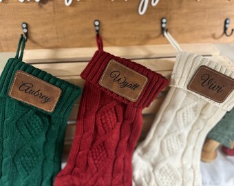 May include: Three cable-knit Christmas stockings in green, red, and cream, each with a brown leather patch bearing a name: Aubrey, Wyatt, and Vivi. The stockings hang from black hooks on a wooden board, perfect for holiday decor.