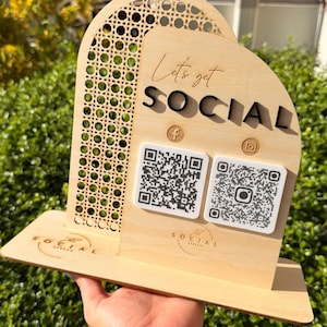 May include: A light-colored wooden sign with an arched top and a cane webbing panel. It features the text "Let's get SOCIAL" in black and script lettering, two square QR code placards, and social media icons for Facebook and Instagram.