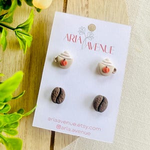 May include: A white card displays four earrings: two coffee cups with whipped cream and pumpkin accents, and two coffee beans. The card reads "ARIA AVENUE" and includes a website and Instagram handle.
