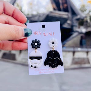 May include: A pair of handmade polymer clay earrings. One earring features a black and white Stormtrooper design, and the other a black Darth Vader design. Each earring is topped with a flower. The earrings are displayed on a white card with the text "ARIA AVENUE".