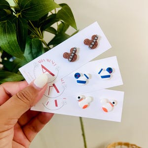 May include: A collection of handmade stud earrings featuring three distinct designs. One pair resembles a brown furry character with a bandolier, another has a blue and white design, and the last pair is white with orange accents.