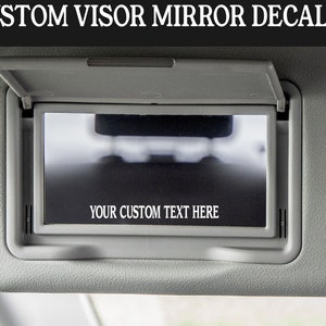 K&ouml;nnte beinhalten: Ein grauer Auto-Sonnenblendenspiegel mit einem schwarzen rechteckigen Aufkleber, auf dem "YOUR CUSTOM TEXT HERE" steht.