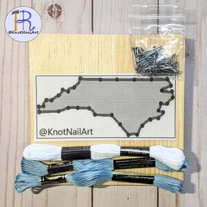 May include: A DIY string art kit featuring the outline of the state of North Carolina. The kit includes a wooden board, a bag of nails, and embroidery floss in white and blue.