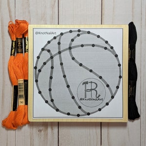 May include: A wooden board with a black and white basketball design. The board has black dots around the outline of the basketball. There are three orange spools of embroidery floss and one black spool of embroidery floss on the left and right sides of the board. The text "@KnotNailArt" is printed on the board.