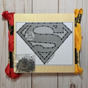May include: A Superman logo nail art kit on a wooden board. The kit includes a printed design, nails, red and yellow embroidery floss. The logo is gray with black dots, and the text "@KnotNailArt" is visible.