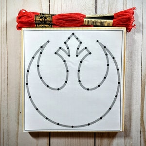 May include: A white wooden frame with a black and gray Rebel Alliance symbol. The symbol is made up of a series of dots that are connected by lines. The frame is surrounded by red embroidery floss.