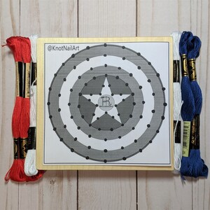 May include: A wooden board with a printed design of Captain America's shield. The shield is gray and white with a red star in the center. The board is surrounded by embroidery floss in red, white, and blue.