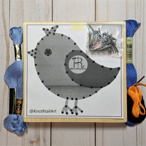 May include: A gray bird-shaped string art kit with black dots for the stitching. The kit includes blue, black, and orange embroidery floss, a bag of nails, and a wooden frame. The frame has the text "@KnotNailArt" printed on it.