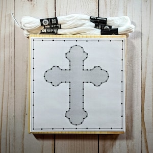 May include: A craft kit featuring a cross design for string art. The kit includes a wooden base with a printed cross outline and pre-drilled holes, along with several spools of white embroidery floss. The wooden base is rectangular with a light wood color.