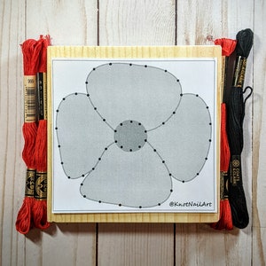 Op de afbeelding: Een houten knutselset met een grijs en wit klaproosontwerp. De set bevat rode en zwarte borduurgaren en de tekst "@KnotNailArt".