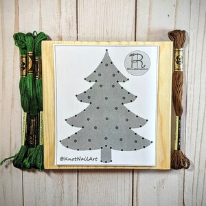 May include: A wooden frame with a grey and white Christmas tree design with black dots. The frame is surrounded by green and brown embroidery floss.