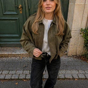 Può includere: Giacca verde oliva con bottoni, indossata sopra un maglione chiaro e una camicia bianca. L'outfit è completato da jeans neri e una cintura nera con fibbia dorata. Le scarpe marroni completano il look.
