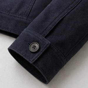 Pu&ograve; includere: Primo piano di una manica di giacca in camoscio blu navy. Il polsino presenta una chiusura con bottoni con un bottone scuro. Le cuciture dettagliano le cuciture e l'asola. La giacca sembra essere realizzata in un materiale morbido e strutturato.
