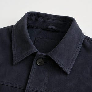 Pu&ograve; includere: Primo piano di una giacca blu navy con colletto classico e chiusura con un solo bottone. La giacca presenta dettagli di cucitura visibili e una piccola tasca. Il tessuto ha un aspetto leggermente usurato.