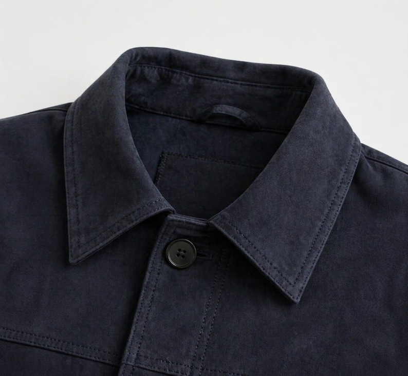 Pu&ograve; includere: Primo piano di una giacca blu navy con colletto classico e chiusura con un solo bottone. La giacca presenta dettagli di cucitura visibili e una piccola tasca. Il tessuto ha un aspetto leggermente usurato.