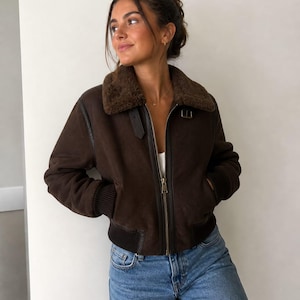 Può includere: Giacca bomber marrone scuro con colletto in montone, chiusura con zip e polsini a coste. La giacca è abbinata a jeans blu. La giacca ha un design classico e un ricco colore marrone.