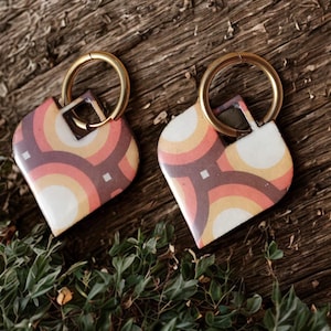 Puede incluir: Dos llaveros en forma de corazón con un diseño retro. Los llaveros presentan un patrón de líneas curvas en tonos morado, naranja y blanco. Cada llavero tiene un anillo dorado para sujetar las llaves.