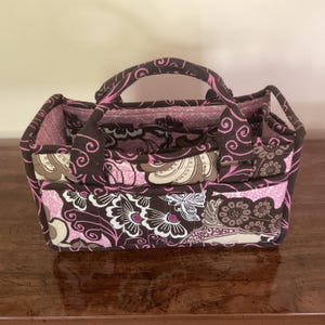 May include: A fabric tote bag with a floral pattern in shades of brown, pink, and white. The bag has a handle and multiple compartments, perfect for organizing items. The bag is approximately 10 inches wide and 7 inches tall.