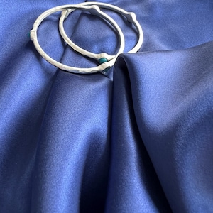 K&ouml;nnte beinhalten: Zwei silberne Armreifen mit t&uuml;rkisfarbenen Steinen, die in das Metall eingefasst sind. Die Armreifen liegen auf einem blauen Satin-Stoff.